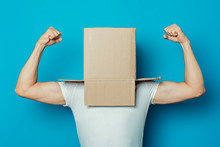 Young Man In A White T-shirt With A Cardboard Box On His Head On A Blue Background. Makes A Hand Gesture Strength And Power, Bodybuilding, Sport