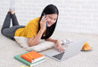 © weedezign - Asian female talk on mobile and use laptop computer to work at home.woman lay down on carpet at brick wall.working online lifestyle