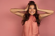 © bestsenny - A dark-haired girl in a pink dress stands on a pink background clasping her hands to her head and smiling, looking at the camera.