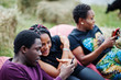 © AS Photo Family - Three african american friends chill, sitting on poufs and using their phones outdoor.
