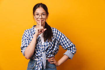 Sticker - Cute teenage girl in glasses posing isolated over yellow wall background showing silence gesture.