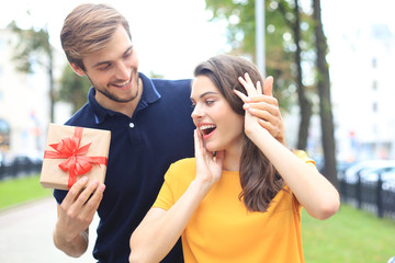 Wall Mural - Astonished excited couple in summer clothes smiling and holding present box together while standing on city street.