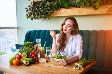 Wall Mural - Young happy woman eating salad in the beautiful interior with green flowers on the background and fresh ingredients on the table. Healthy food concept