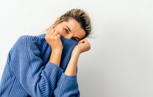 Image Of Beautiful Smiling Young Woman Pulling Her Blue Trendy Sweater Over Head Having Fun. Joyful Female With Tied Hair In Topknot Being Childish Disappearing In Her Clothes Looking To The Camera