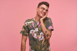 Indoor portrait of cute shy young guy with palm on his cheek, wearing glasses and flowered t-shirt, looking aside with blushing smile, standing over pink background