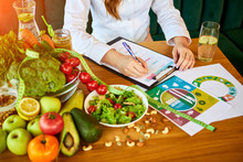 Woman Dietitian In Medical Uniform With Tape Measure Working On A Diet Plan Sitting With Different Healthy Food Ingredients In The Green Office On Background. Weight Loss And Right Nutrition Concept