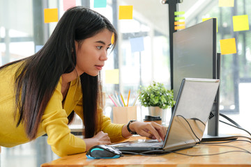 young asian female programmer in a yellow shirt stands down to use her laptop and pc in the office.
