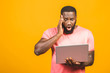 Young surprised african man standing and using laptop computer isolated over yellow background.