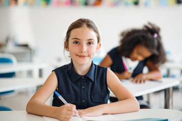 Wall Mural - A small school girl sitting at the desk in classroom, looking at camera.