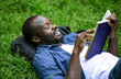 © proimagecontent - Joyful African-american student is carried away by reading lying on a campus park lawn