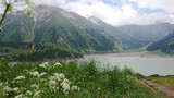 Fototapeta Natura - Big Almaty lake located in the mountains of Kazakhstan. It offers views of green grass, flowers, lake, rocks, large mountains and the sky in the clouds. Mountain lake with blue water.