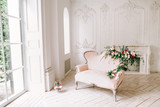Fototapeta Kwiaty - vintage sofa of soft pink color, decorated with flowers and greens, stands in a classic room on a white wooden floor surrounded by lighted candles in glass candlesticks near  large window and curtains