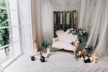 vintage sofa of soft pink color, decorated with flowers and greens, stands in a classic room on a white wooden floor surrounded by lighted candles in glass candlesticks near  large window and curtains