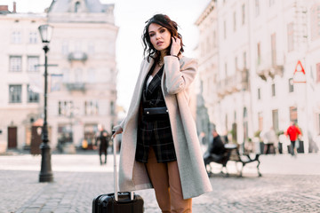 Pretty young woman with short dark hair in casual stylish clothes traveling with a wheeled suitcase, walking at the street of old European city