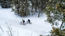 Aerial View Of Two Mountain Bikers Riding On Road In Forest Outdoors In Winter.