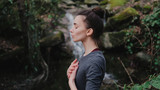 Fototapeta Zwierzęta - Young woman practicing breathing yoga pranayama outdoors in moss forest on background of waterfall. Unity with nature concept.
