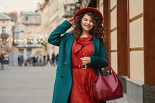 Happy Smiling Fashionable Curvy Woman Wearing Trendy Autumn Outfit: Orange Hat, Snakeskin Print Dress, Belt, Green Coat, Holding Red Wicker Leather Bag, Posing In Street Of European City. Copy Space