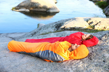 Wall Mural - Young couple resting in sleeping bags on cliff near lake