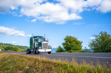 Classic powerful green big rig bonnet semi truck tractor running on summer road with cloud sky