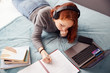 © Monkey Business - Looking Down On Female College Student Wearing Headphones Lying On Bed Working On Laptop