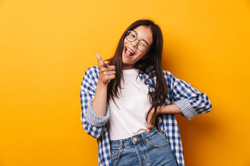 Sticker - Smiling emotional young cute teenage girl in glasses posing isolated over yellow wall background pointing to you.