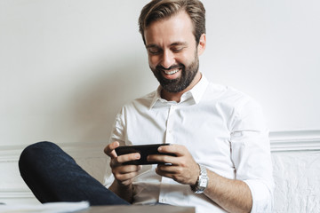 Canvas Print - Image of successful laughing businessman playing video game on cellphone while sitting at table in office