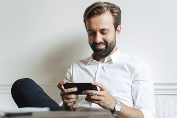 Poster - Image of successful smiling businessman playing video game on cellphone while sitting at table in office