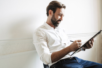 Canvas Print - Image of bearded concentrated businessman holding clipboard and writing down notes while sitting on chair in office