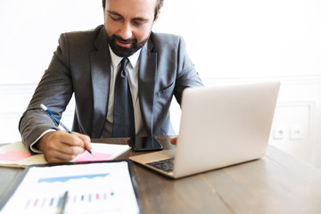 Sticker - Image of concentrated confident businessman working at laptop and cellphone in office while writing down notes