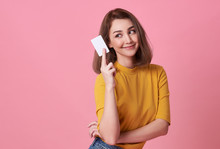 Portrait Of A Young Woman In Yellow Shirt Showing Credit Card And Looking Away At Copy Space Isolated Over Pink Background.