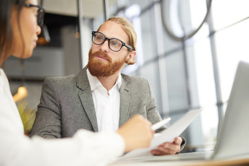 Wall Mural - Serious bearded businessman in eyeglasses holding document and listening to his partner during business meeting at office