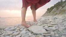 Close-up Slow Motion Following Behind The Legs Of A Barefoot Girl In A Red Dress Fluttering In The Wind At Sunset Walking Crouching Along The Stones Of The Sea Shore. Light Moments Of Tidelessness