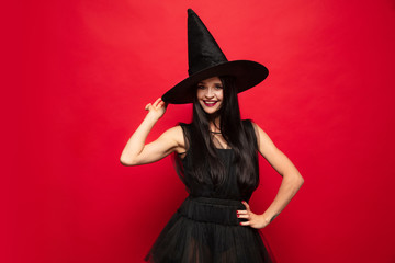 Wall Mural - Young brunette woman in black hat and costume on red background. Attractive caucasian female model. Halloween, black friday, cyber monday, sales, autumn concept. Copyspace. Dancing, posing charming.