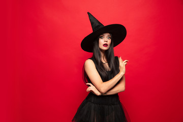 Wall Mural - Young brunette woman in black hat and costume on red background. Attractive caucasian female model. Halloween, black friday, cyber monday, sales, autumn concept. Copyspace. Pointing up.