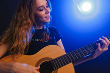 Positive Attractive Young Woman Musician Play On A Guitar On Scene In Night Club Over Dark Background With Flash Lights
