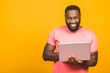 Young smiling african american man standing and using laptop computer isolated over yellow background.