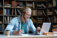 Smiling Happy Young Man Wear Wireless Headset Look At Laptop Screen Make Notes Study E Learning In Library Watch Webinar Training Online Course Video Call, Distance Education, Skype Teaching Concept