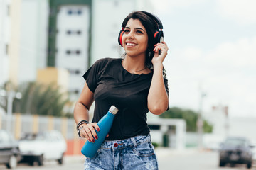 Beautiful young woman using headphone listening to music and drinking water outdoors