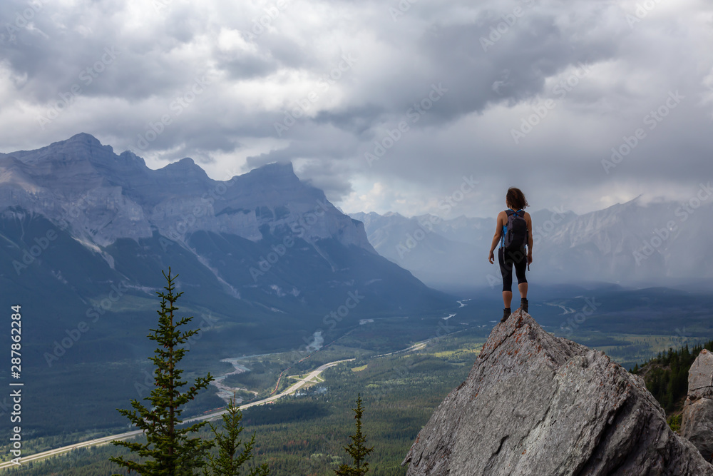 Adventurous Caucasian Girl is hiking up a rocky mountain during a cloudy and rainy day. Taken from Mt Lady MacDonald, Canmore, Alberta, Canada. - obrazy, fototapety, plakaty 