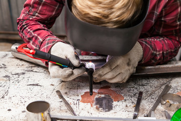 Wall Mural - Young guy welder in a checkered red shirt welds a stainless steel pipe using agronomic welding to protect his eyes with a mask in an iron workshop. Modern welding methods.