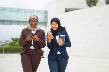 Joyful office female friends with smartphones chatting outside. Muslim business women in hijabs walking outdoors, using mobile phones, talking, laughing. Office friendship concept