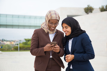 Wall Mural - Excited happy office employees looking at smartphone screen outside. Muslim business women in hijabs standing outdoors, using mobile phones together and laughing. Digital technology concept