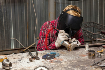 Wall Mural - Young guy welder in a checkered red shirt welds a stainless steel pipe using agronomic welding to protect his eyes with a mask in an iron workshop. Modern welding methods.