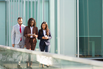 Successful business team going along outdoor urban glass wall. Business man and women walking outside, using tablet, drinking coffee, talking. Teamwork concept