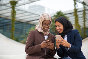 Wall Mural - Happy office colleagues chatting on smartphones and sharing news. Muslim business women in hijabs sitting outside, using mobile phones together, texting messages. Communication concept