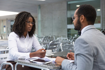 Serious business colleagues discussing ongoing project over cup of coffee. Business man and woman meeting at table with gadgets and papers in street cafe and talking. Business conversation concept