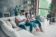 © deagreez - Nice attractive lovely cheerful cheery friendly adorable healthy family wearing casual white t-shirts jeans sitting on sofa using 5g app browsing at industrial loft style interior living-room