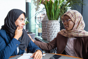 Wall Mural - Serious African American employee supporting colleague. Muslim businesswomen chatting in coffee shop. Corporate relationship concept