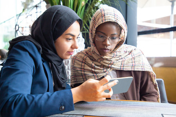 Wall Mural - Focused female colleagues watching project presentation on phone together. Muslim business women in hijabs sitting in cafe, using smartphones, looking at screen. Working in cafe concept