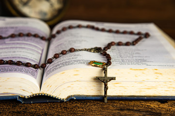 Wall Mural - Closeup open holy bible and beautiful Christian rosary beads on top with blurred vintage alarm clock on wooden background.